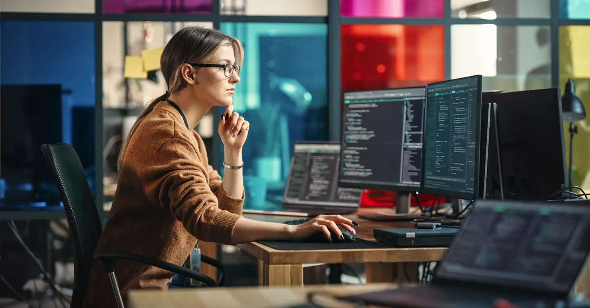 Individual sitting at a desk in a modern office, working on a multi‑monitor setup displaying lines of code and development tools, with colorful glass panels in the background.