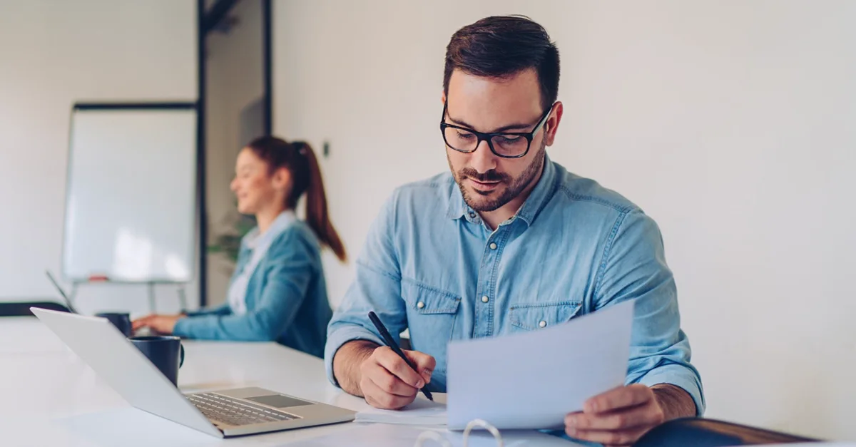 Two individuals working at a desk in an office setting, with one person reviewing papers and writing, and another person working on a laptop in the background.