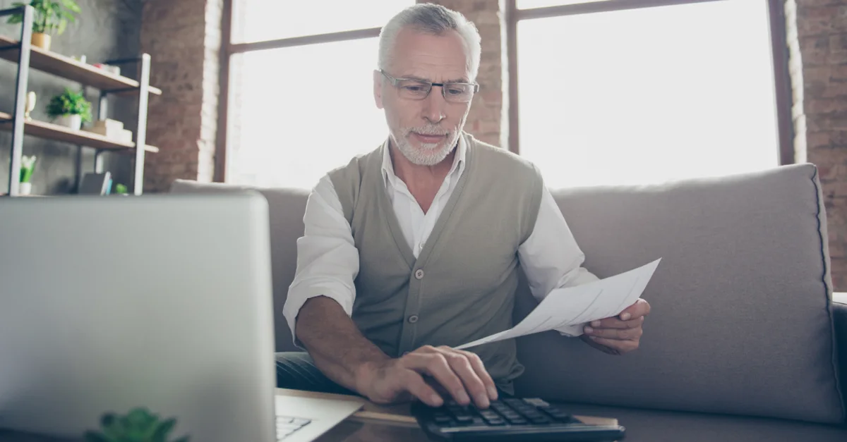 Individual sitting on a sofa working with a laptop, holding documents in one hand and using a calculator on the table in front, with large windows in the background.
