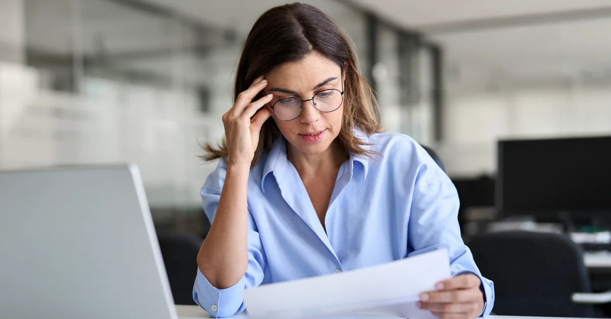 Individual seated at a desk in an office setting, holding and reviewing documents while working on a laptop, with additional papers and office equipment visible in the background.