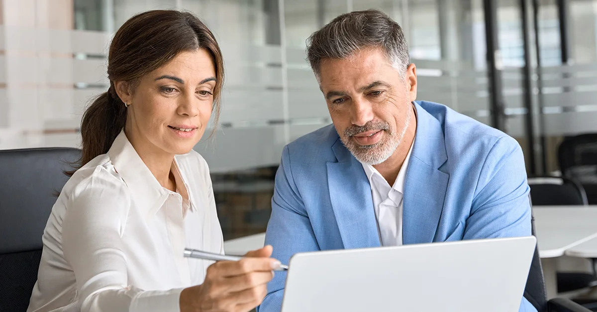 Two individuals sitting closely at a desk in a modern office setting, looking at a laptop screen while one person points with a pen.