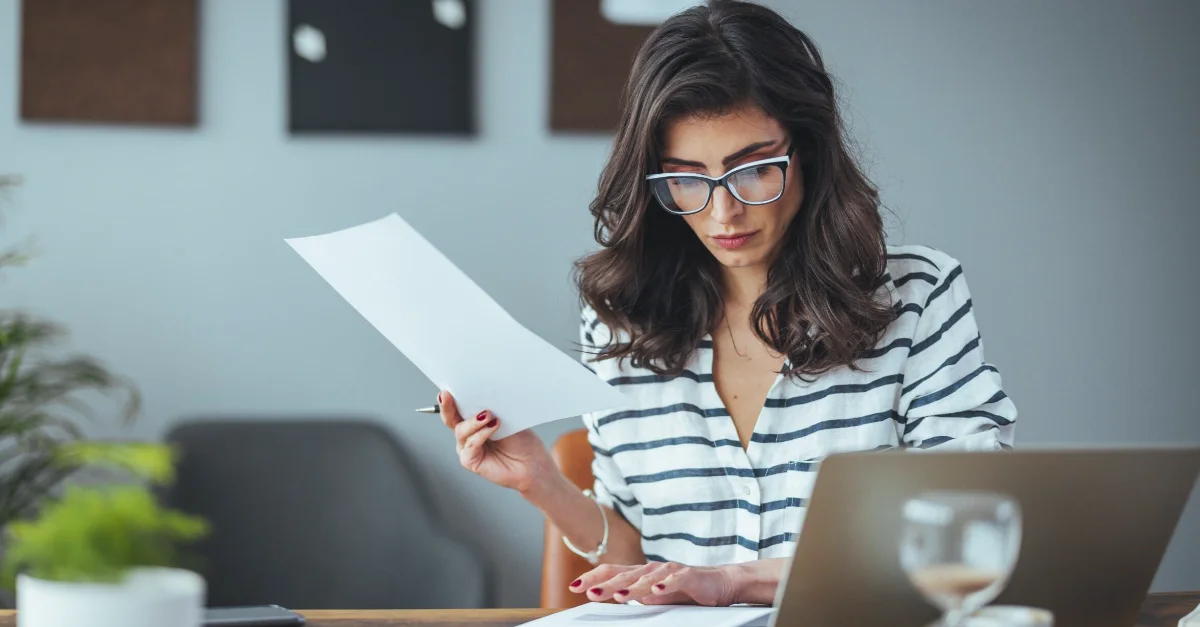 Individual sitting at a wooden desk holding a sheet of paper while working on a laptop, with documents, a notebook, and a glass of water on the table in a home or office workspace.