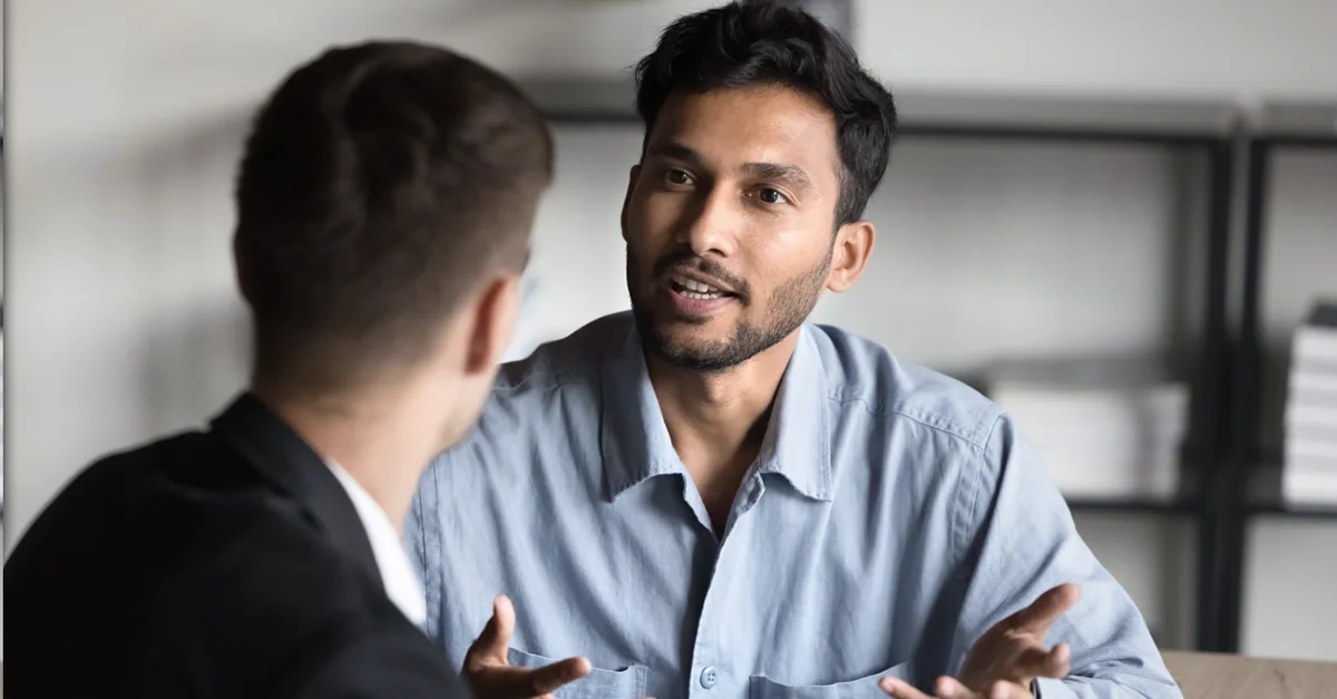 Two individuals seated at a desk in an office setting, engaged in a conversation, with one person using expressive hand gestures while speaking.