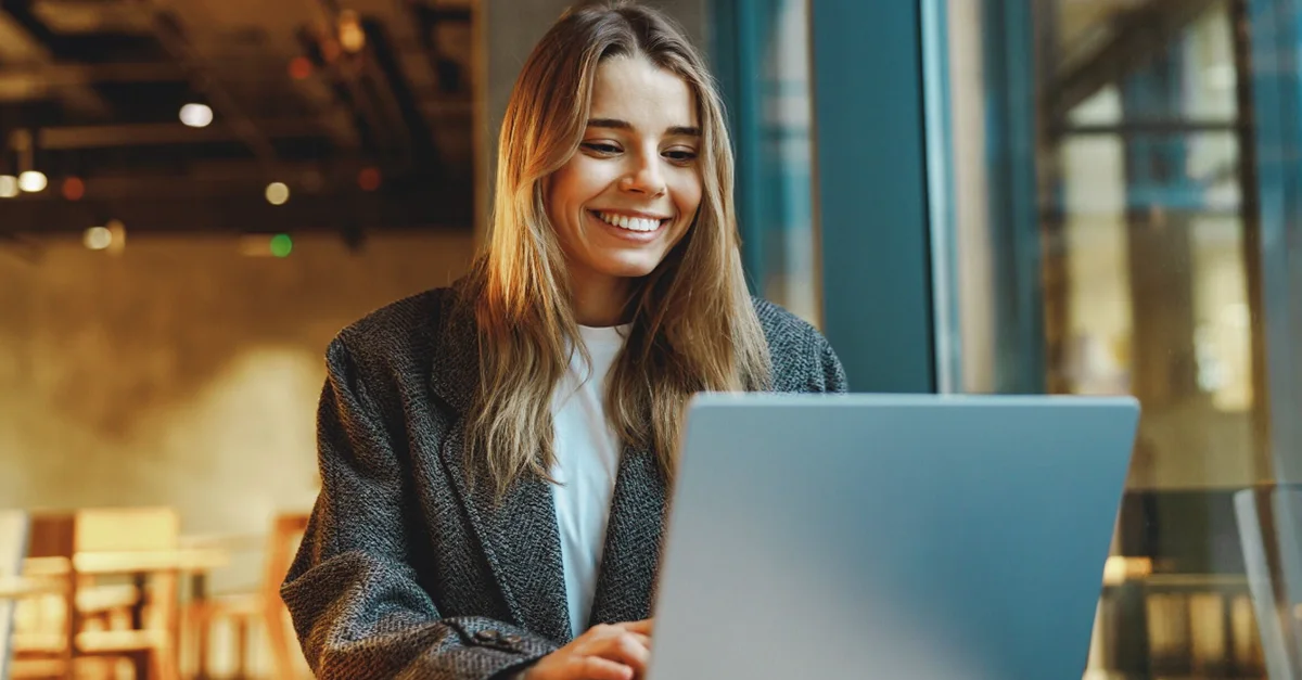A person sitting indoors at a table using a laptop, with large windows and a warm, modern café-style background.
