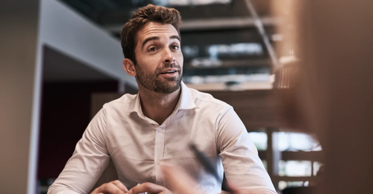 A person seated at a table indoors, holding an object in the hands, with another person partially visible in the foreground and a modern office environment in the background.