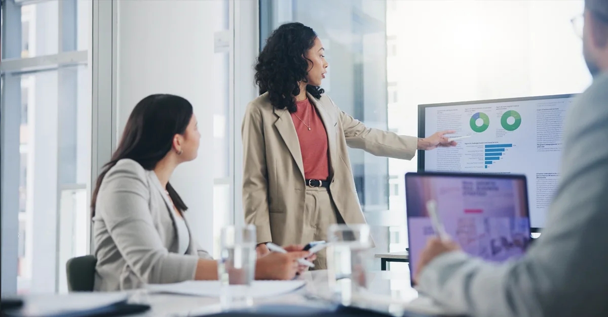 A person standing near a large display screen in a modern office pointing to charts and graphs, while two other people seated at a table look toward the screen. Laptops, glasses of water, and office furniture are also visible.
