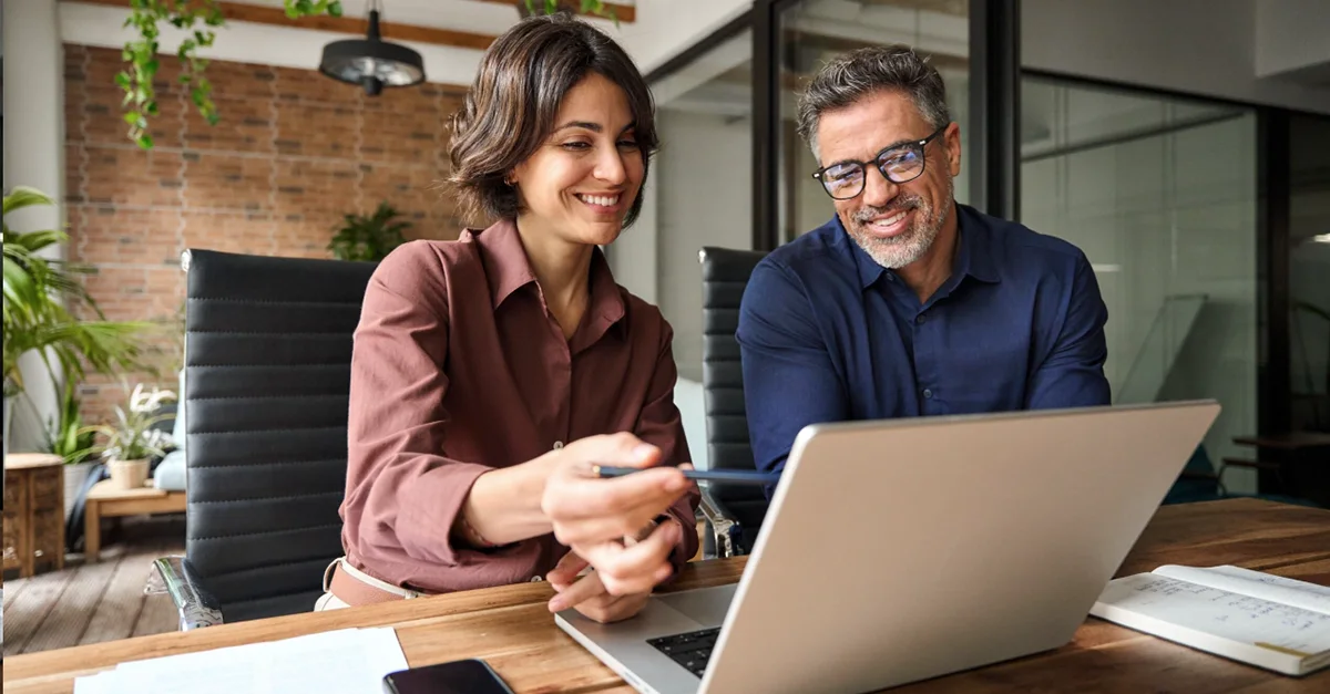 Two people sitting at a wooden table in a modern office, looking at a laptop while one person points at the screen. Papers, a smartphone, and notebooks are placed on the table, and the background shows glass partitions and indoor plants.