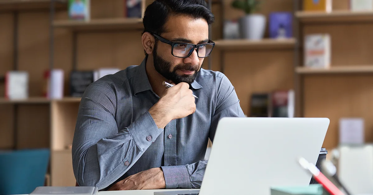 An individual seated at a desk working on a laptop, resting a hand near the chin, with shelves of books and decorative items visible in a softly lit home or office workspace.