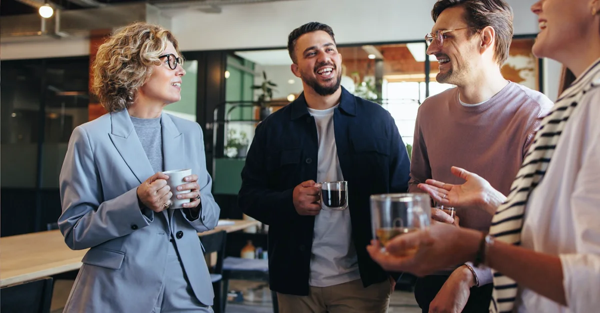 A group of colleagues standing together in a modern office break area, holding coffee cups and having an informal conversation.
