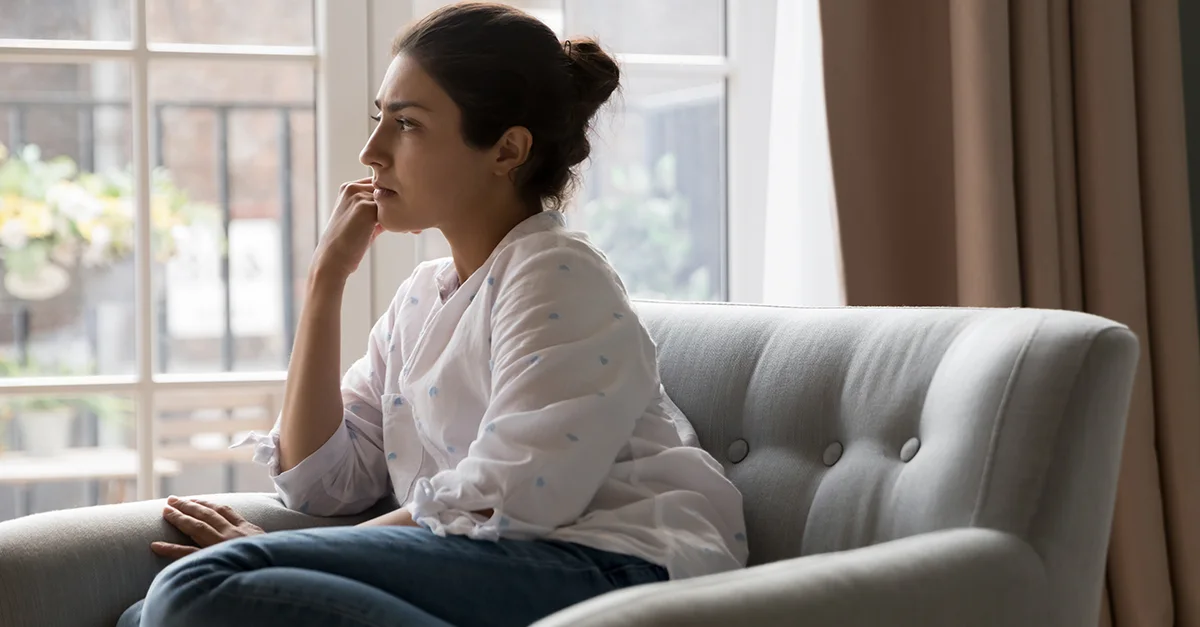 Individual seated on a sofa beside a laptop, resting a hand near the face in a living room setting.