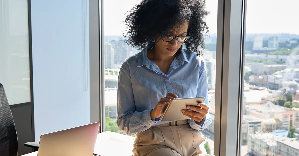 Individual seated at a desk holding a smartphone while looking at a tablet placed on the table.