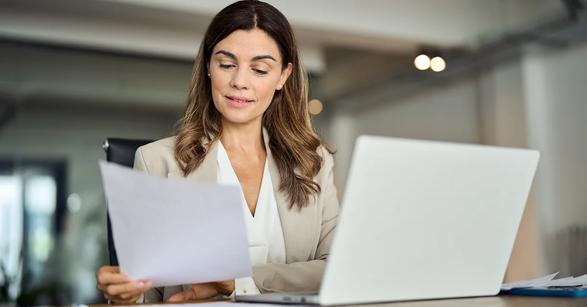 Individual seated at a desk reviewing printed documents while working on a laptop in an office setting.