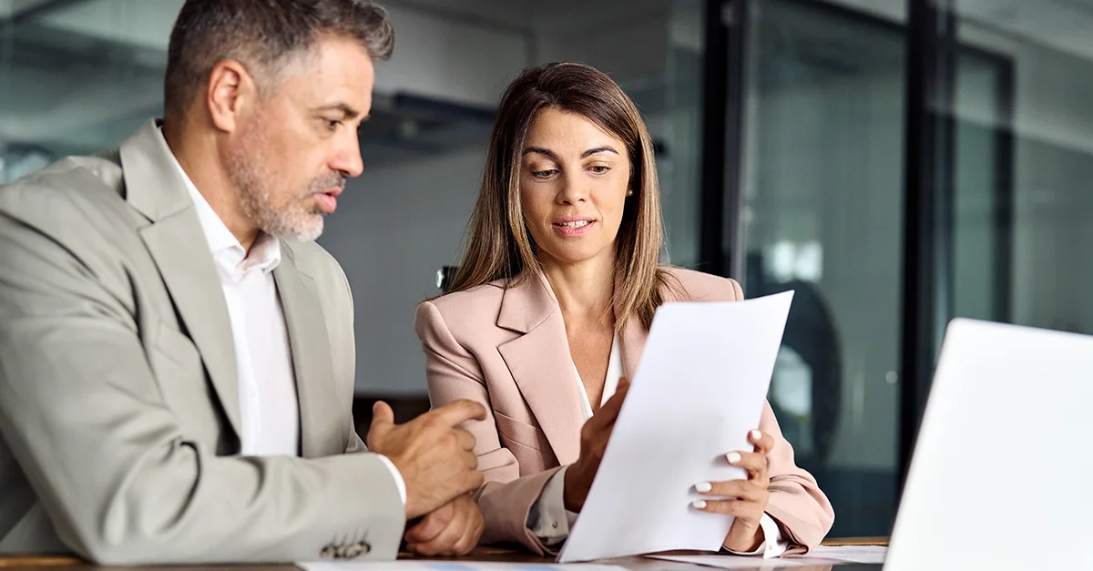 Two individuals standing together in an office, reviewing a printed document.