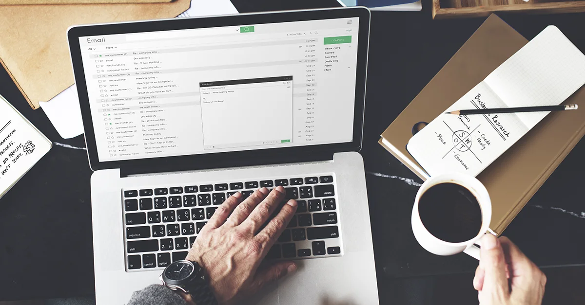Hands typing on a laptop keyboard beside a notebook and a cup of coffee on a desk.
