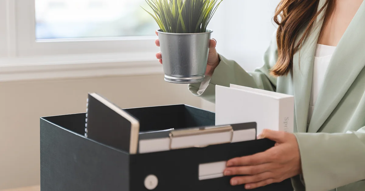 Individual holding a small plant while organizing folders in a storage box on a desk.