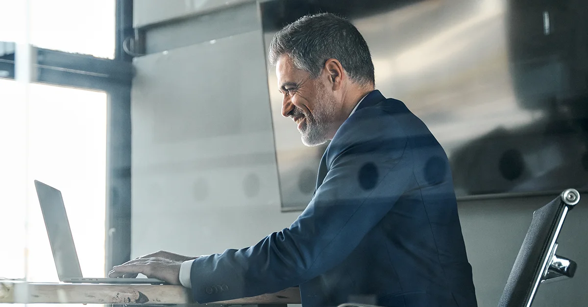 Individual seated at a desk using a laptop near a window in a modern workspace.