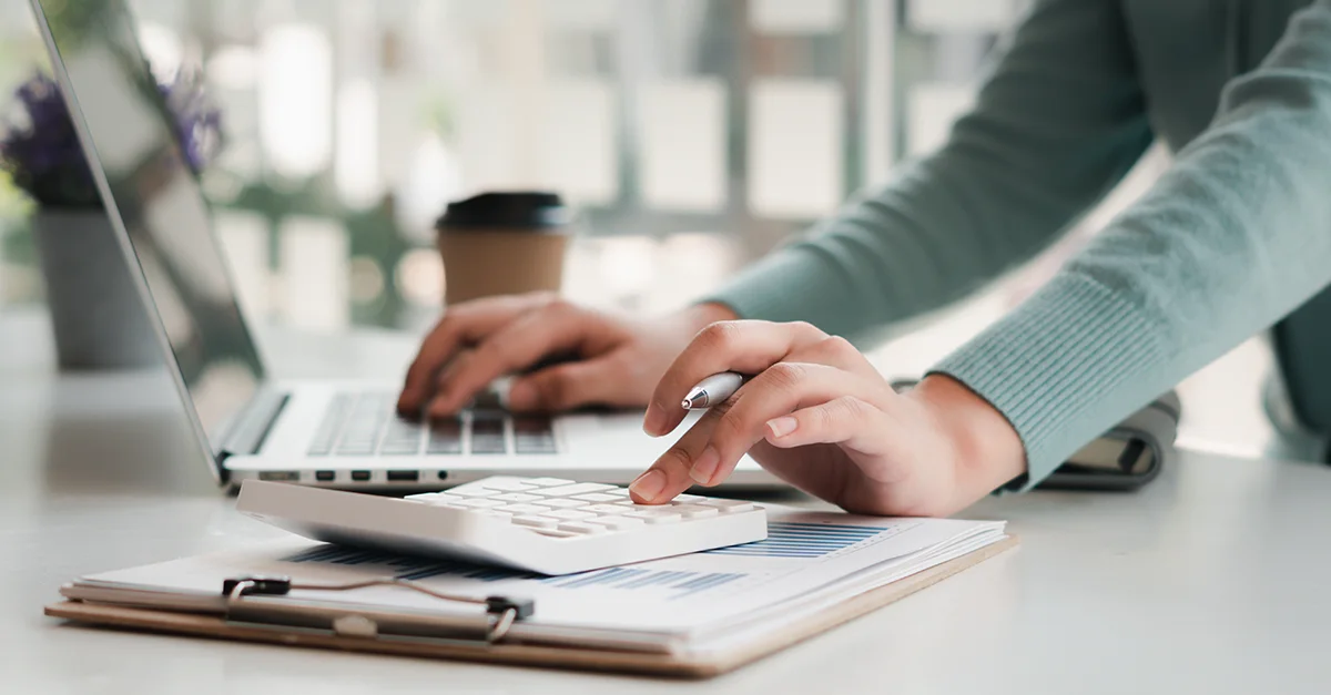 Hands using a calculator at a desk with a laptop, clipboard papers, and a takeaway coffee