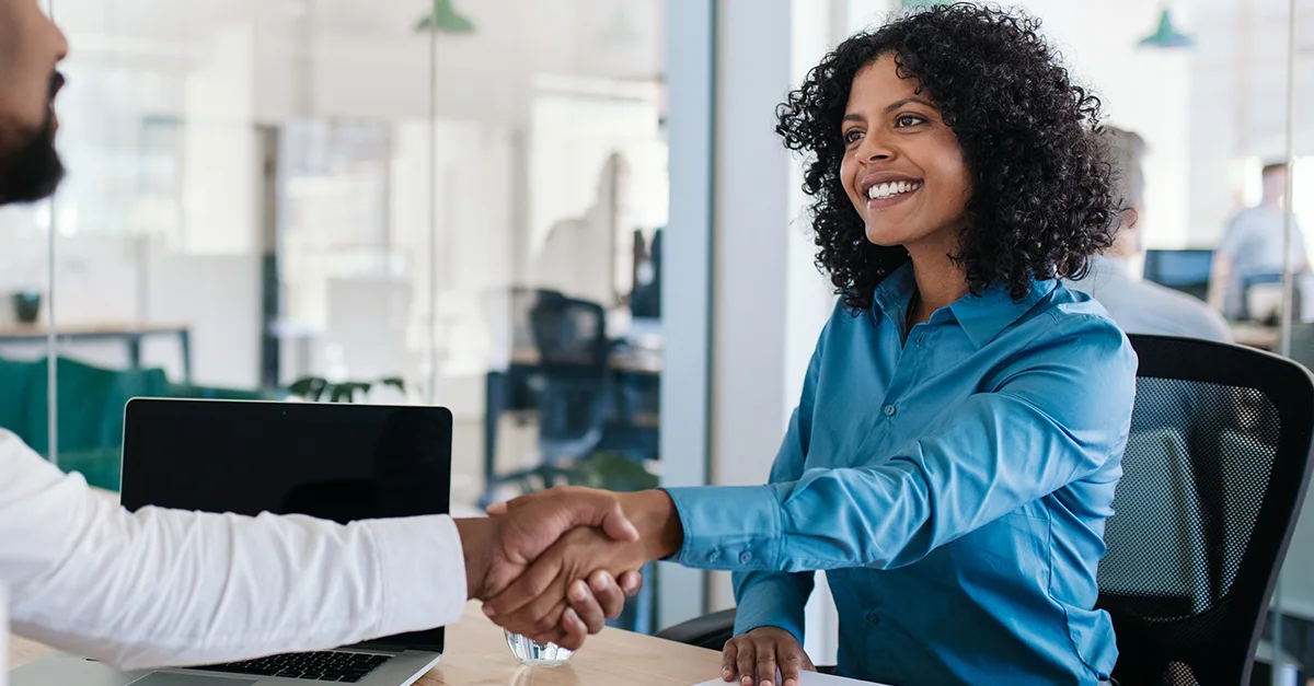 Two people shaking hands across a desk in a modern office with a laptop in the background