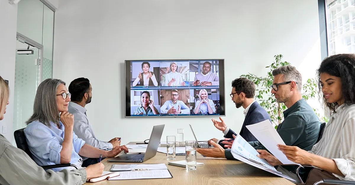 People seated at a conference table while participating in a video conference displayed on a large screen in a meeting room.