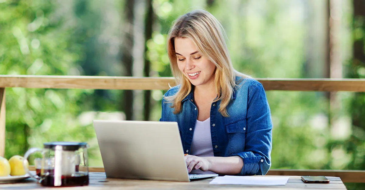 Person working on a laptop at a wooden table outdoors, with a coffee pot and lemons nearby and trees in the background.