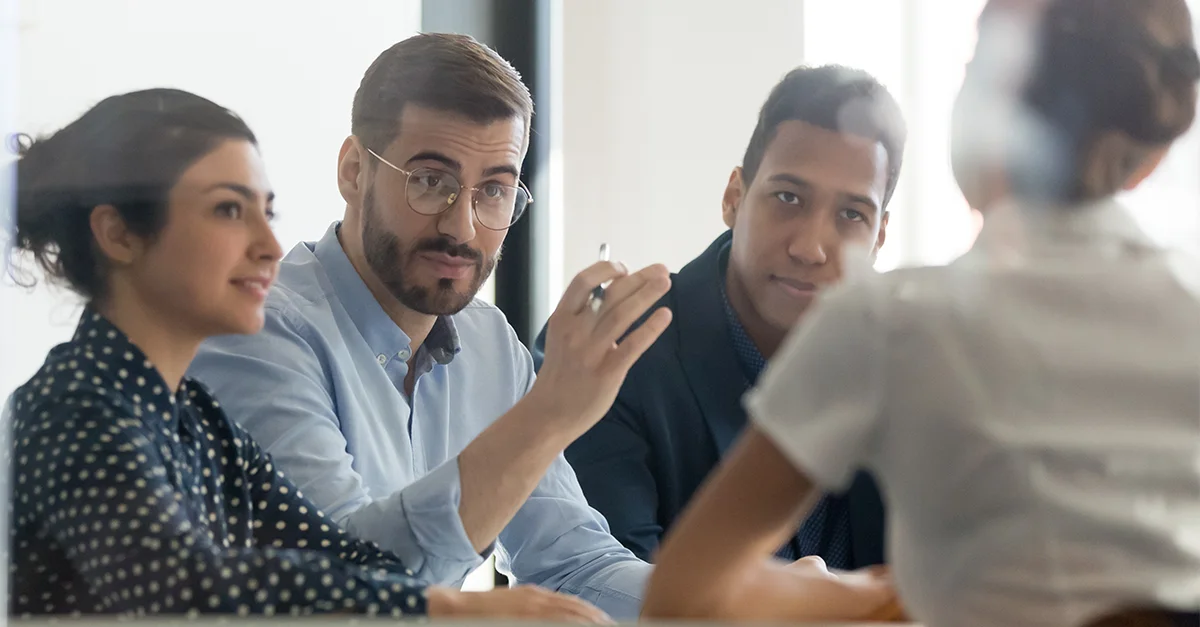 Group of professionals seated at a table in an office setting, engaged in a collaborative discussion, with one person gesturing while speaking.