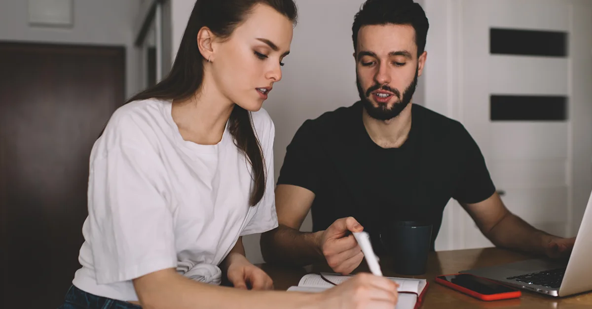 Two people seated at a table at home, reviewing a notebook and laptop while one person writes notes, representing financial planning or collaborative work.