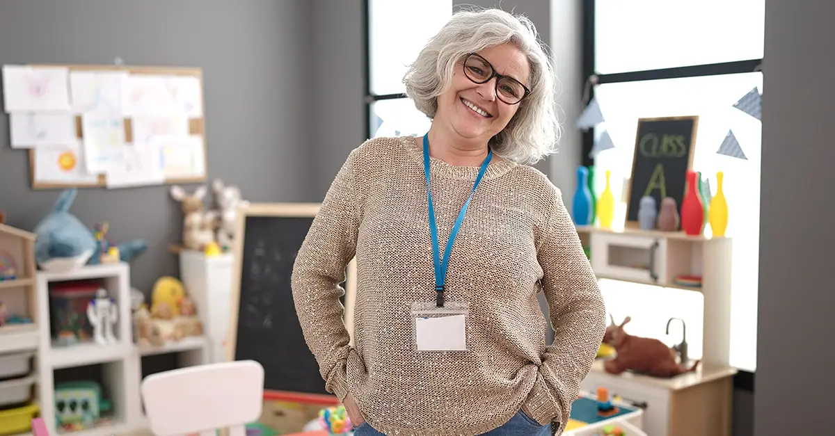 Person standing in a classroom or learning space with shelves of educational materials in the background, wearing a lanyard and casual professional clothing.
