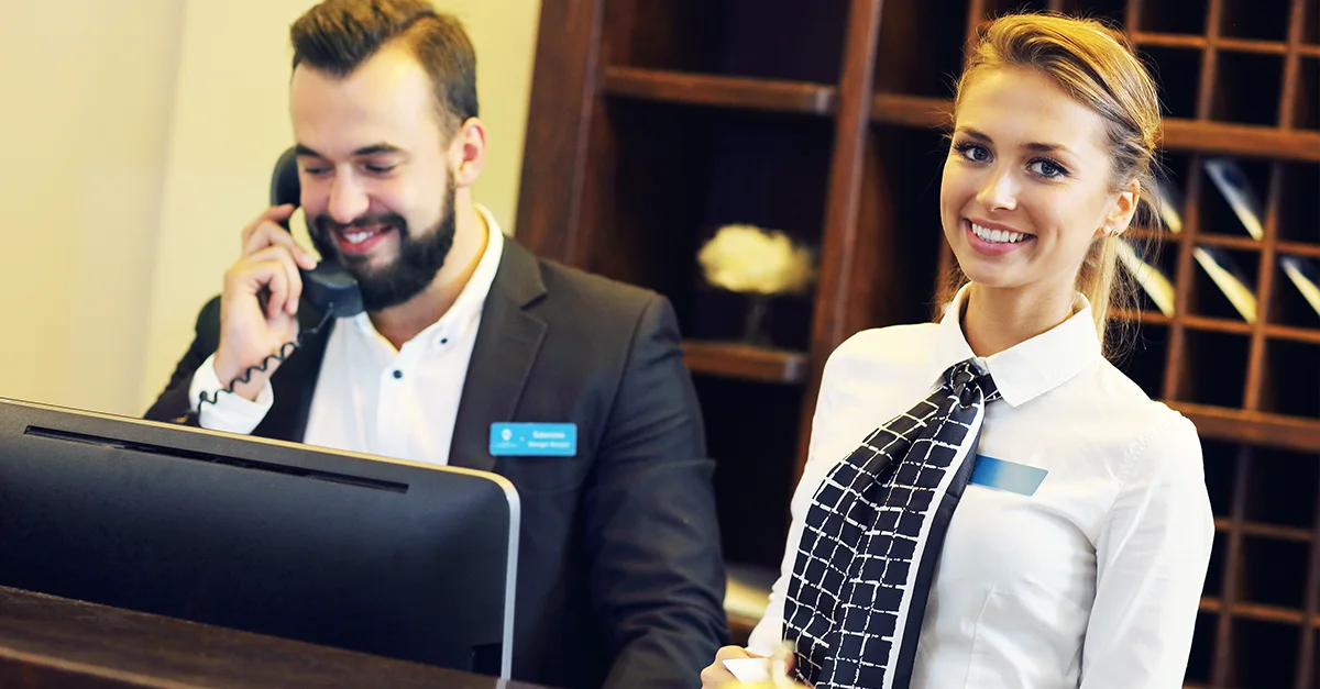Two hospitality professionals standing at a front desk, one using a computer and phone, with a reception area and shelving visible in the background.