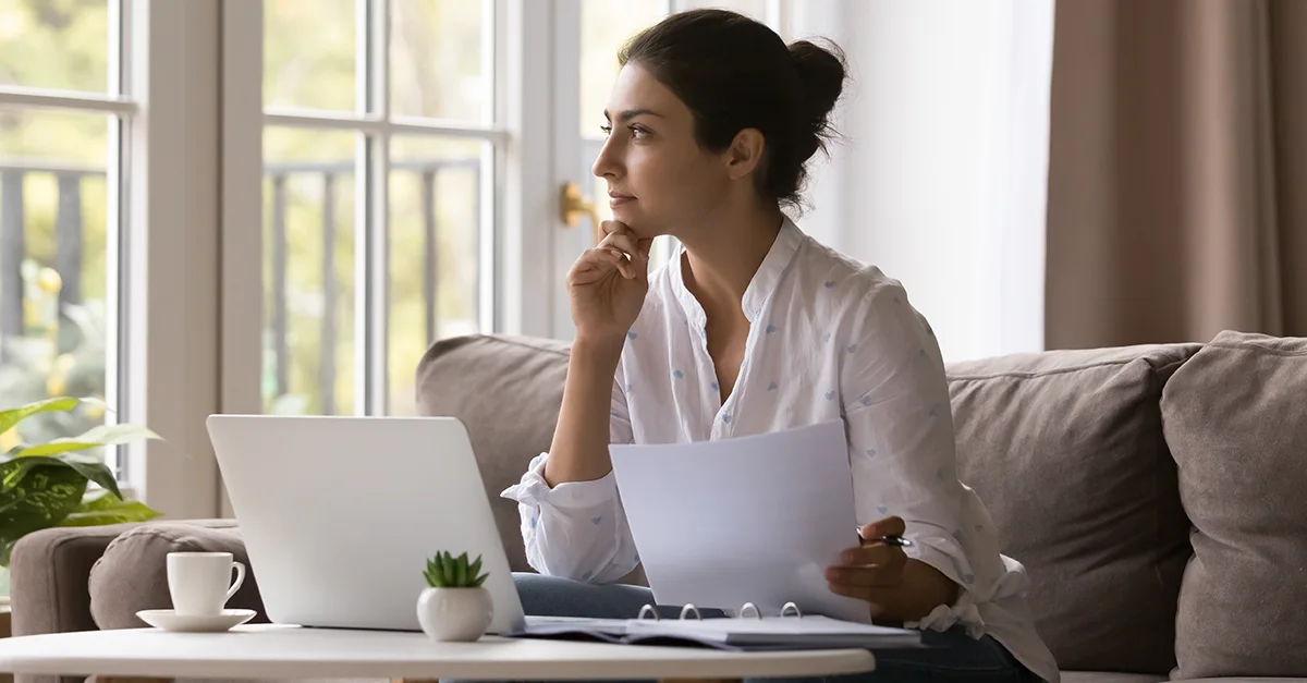 Person seated on a sofa at home, holding papers and looking out a window while working on a laptop on a coffee table, with natural daylight illuminating the room.