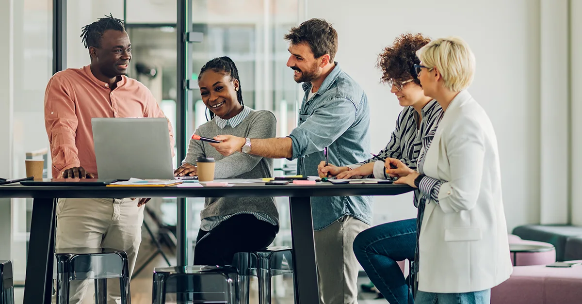 Coworkers gathered around a high table, collaborating on a laptop and discussing ideas in a modern office space.