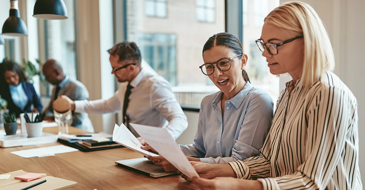 Group of professionals seated at a conference table in a bright office, reviewing printed documents and working with laptops during a meeting.