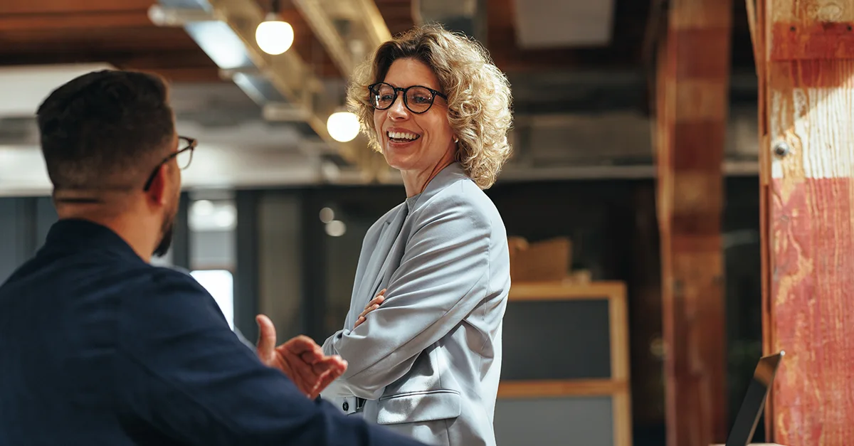 Two coworkers standing and conversing in a modern office interior, with desks, shelving, and overhead lighting visible in the background.