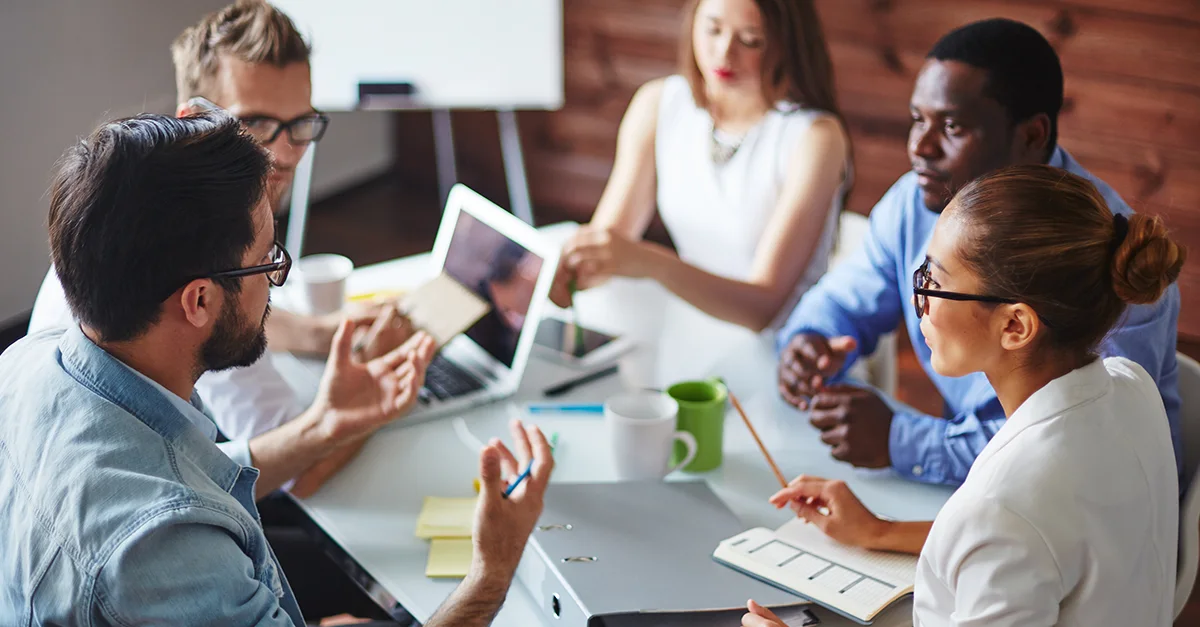 Group of coworkers seated around a table in an office, discussing ideas while reviewing a tablet, notebooks, and printed materials during a collaborative meeting.