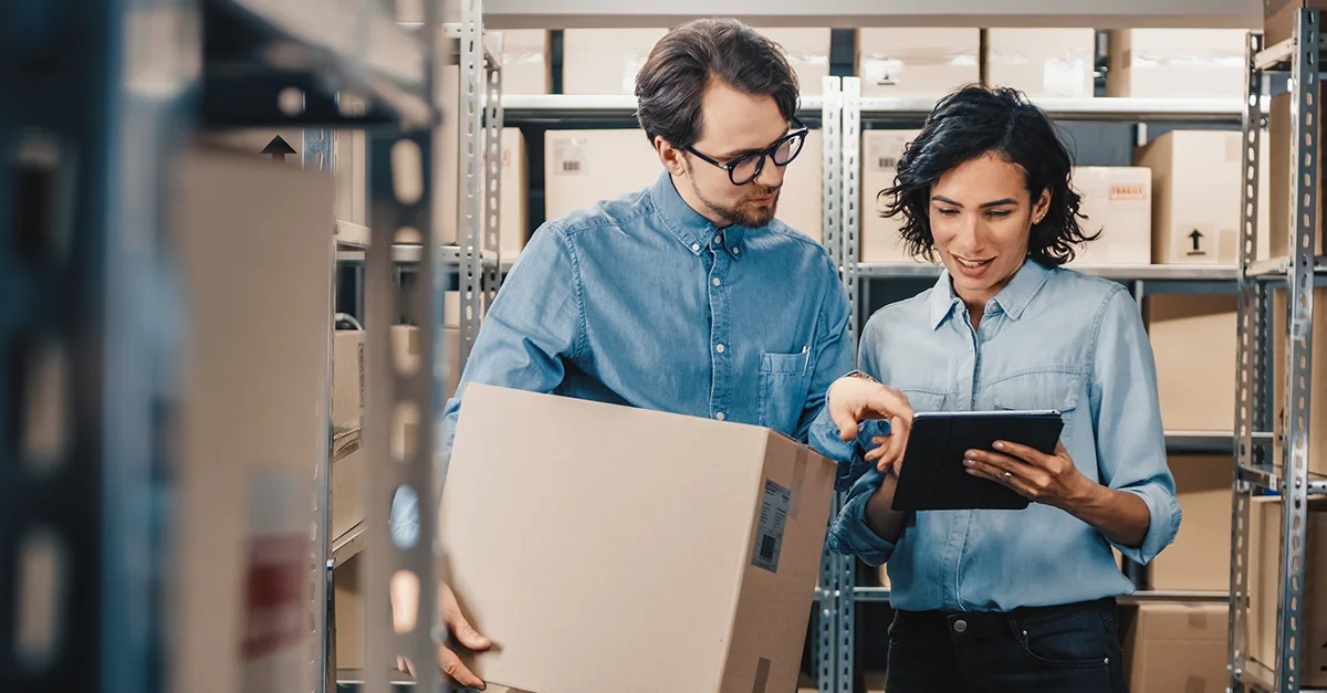 Two coworkers standing in a warehouse aisle with shelving and boxes, reviewing inventory information on a tablet while holding a cardboard box.
