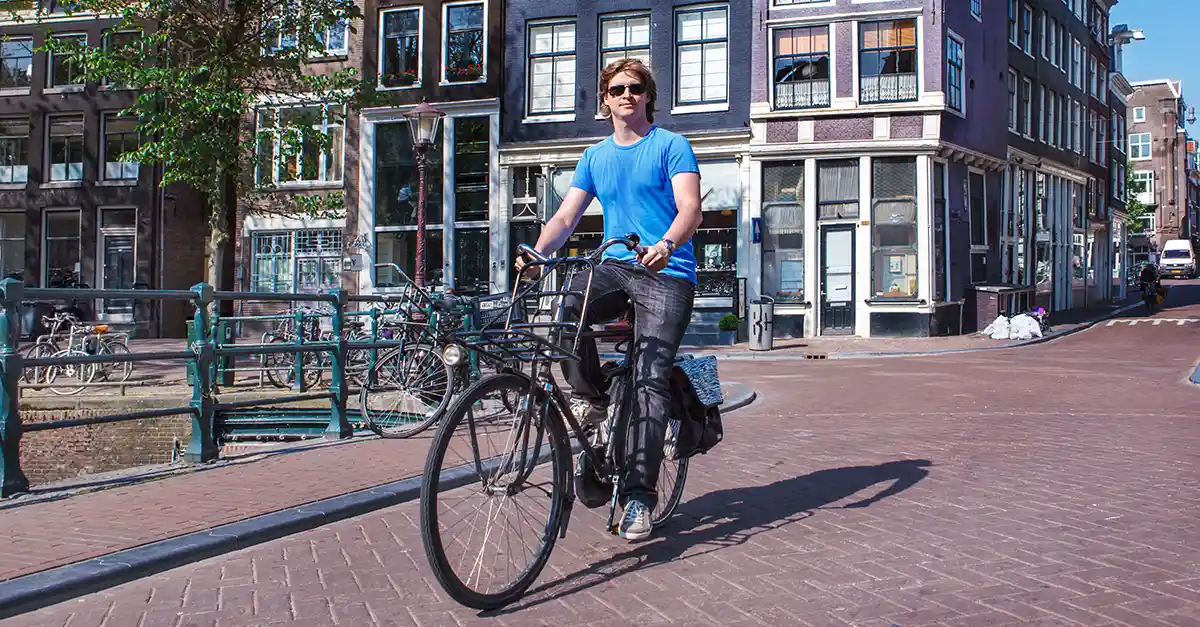 Person riding a bicycle along a paved city street beside canal-side buildings on a sunny day.