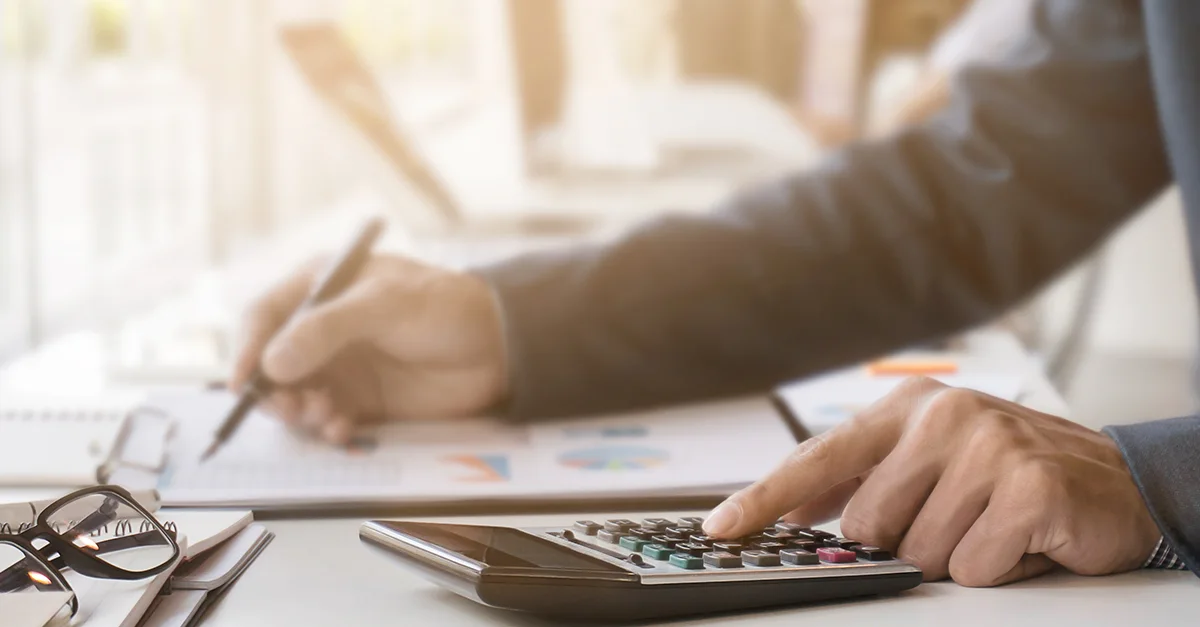 Close-up of hands using a calculator and pen while reviewing printed charts and documents on a desk, with eyeglasses nearby, representing financial analysis or budgeting work.