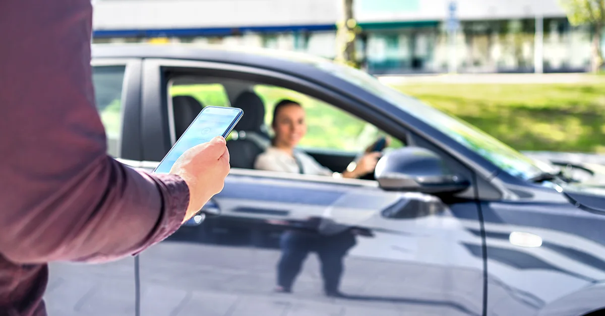 Person holding a smartphone beside a parked car while the driver sits inside, suggesting a mobile app–based interaction or roadside service scenario.