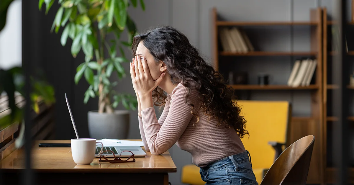 Person seated at a desk with an open laptop, leaning forward with hands covering the face, with a coffee mug, eyeglasses, bookshelves, and indoor plants visible in a home office setting.