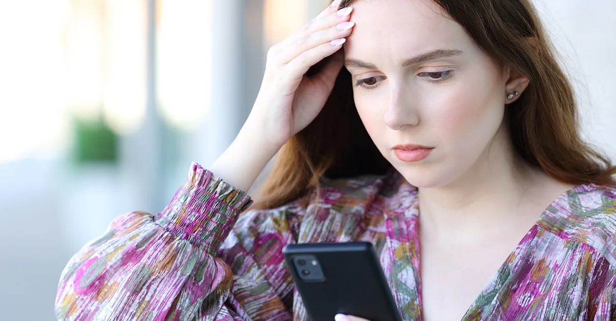 Person holding a smartphone indoors while touching the forehead with one hand, wearing a patterned top, with a softly blurred background.