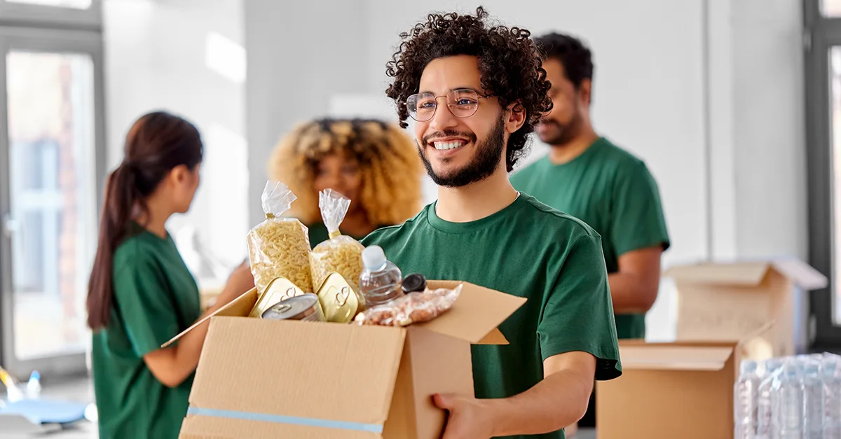 Group of people standing indoors, holding and packing cardboard boxes filled with food items and supplies, with additional boxes stacked in the background, suggesting a donation or volunteering activity.