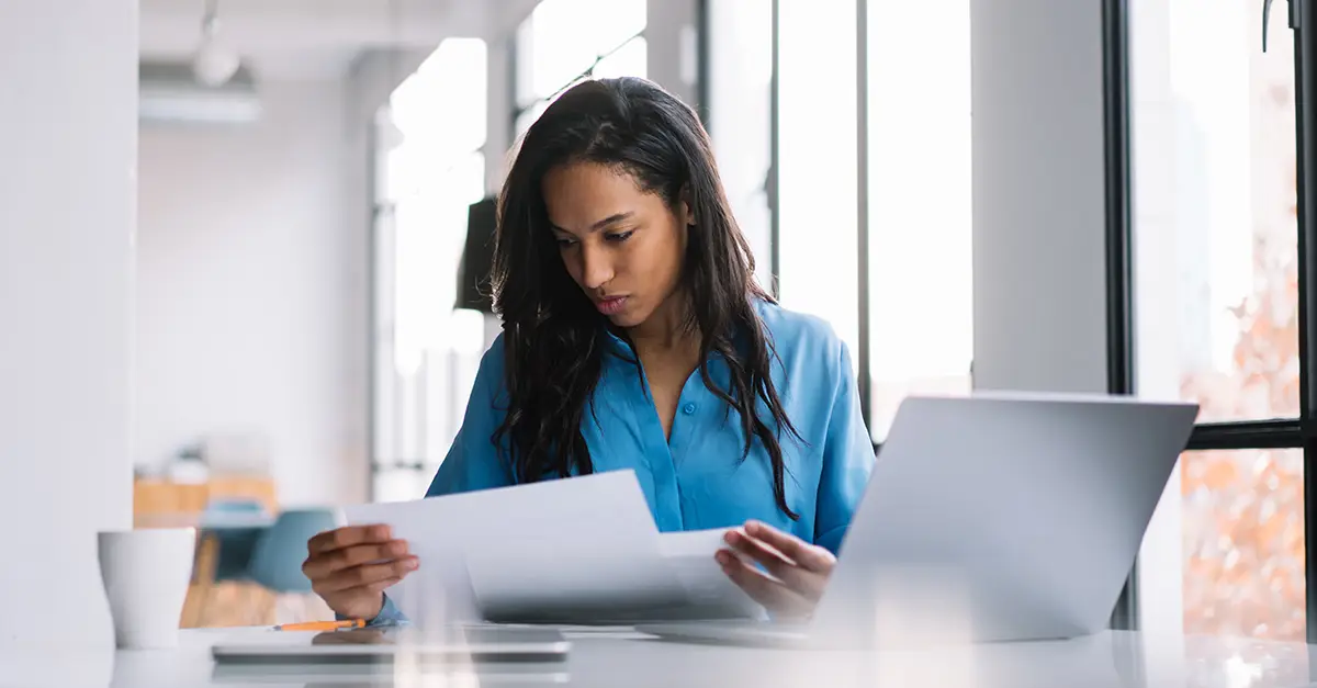 Person reviewing paperwork at a desk with a laptop in a bright office