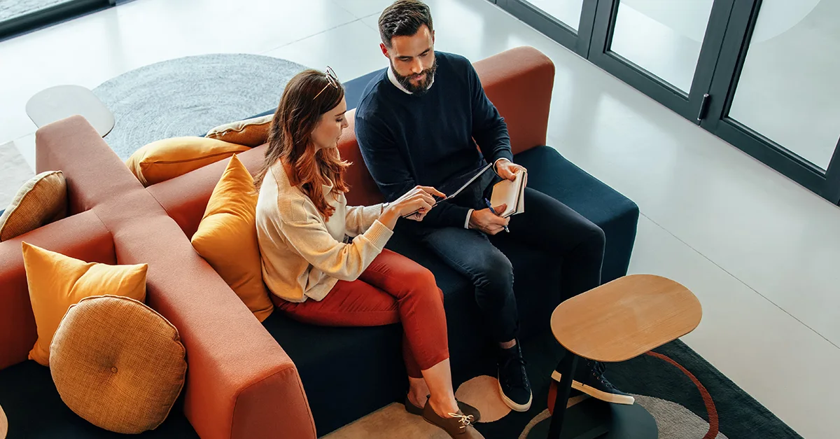 Two people seated on a sofa indoors, looking at a tablet together, with one person holding a credit card and a small table nearby, suggesting online shopping or financial planning.