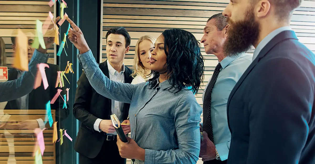 Group of coworkers gathered around a glass board covered with colorful sticky notes, collaborating and pointing at ideas during a brainstorming session in a modern office.