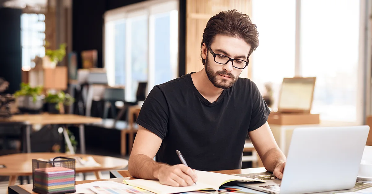 Person seated at a desk in a modern office, writing notes on paper while a laptop sits open nearby, with shelves, plants, and office equipment in the background.
