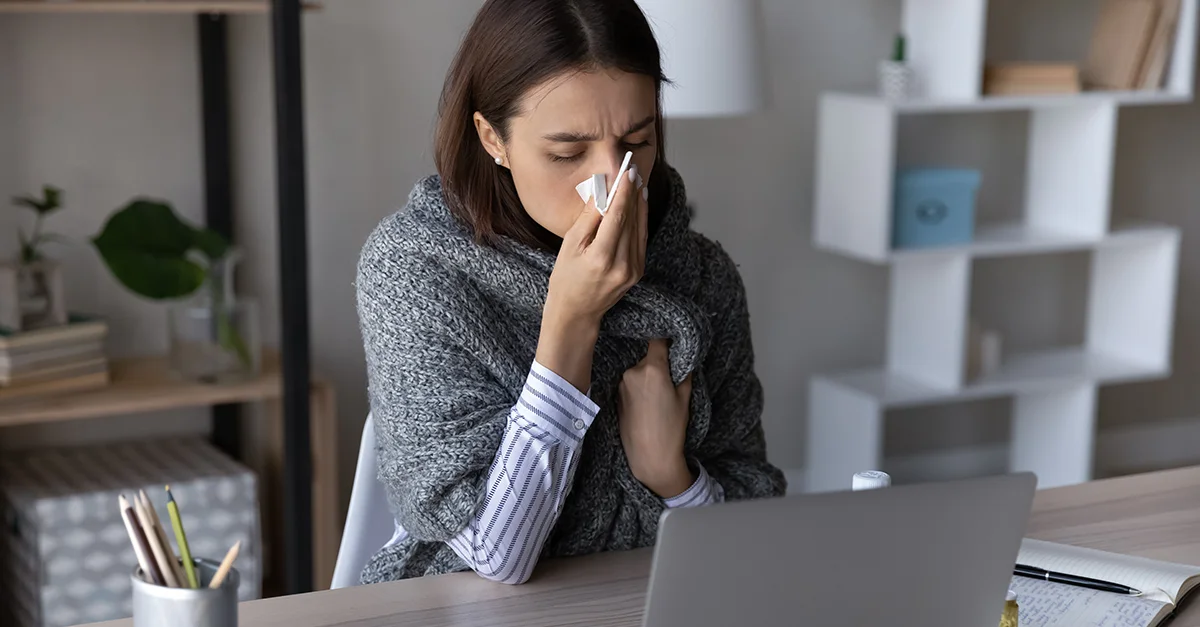 Person seated at a desk using a laptop while holding a tissue to the nose, with a blanket draped over the shoulders and shelves and plants visible in a home interior background.