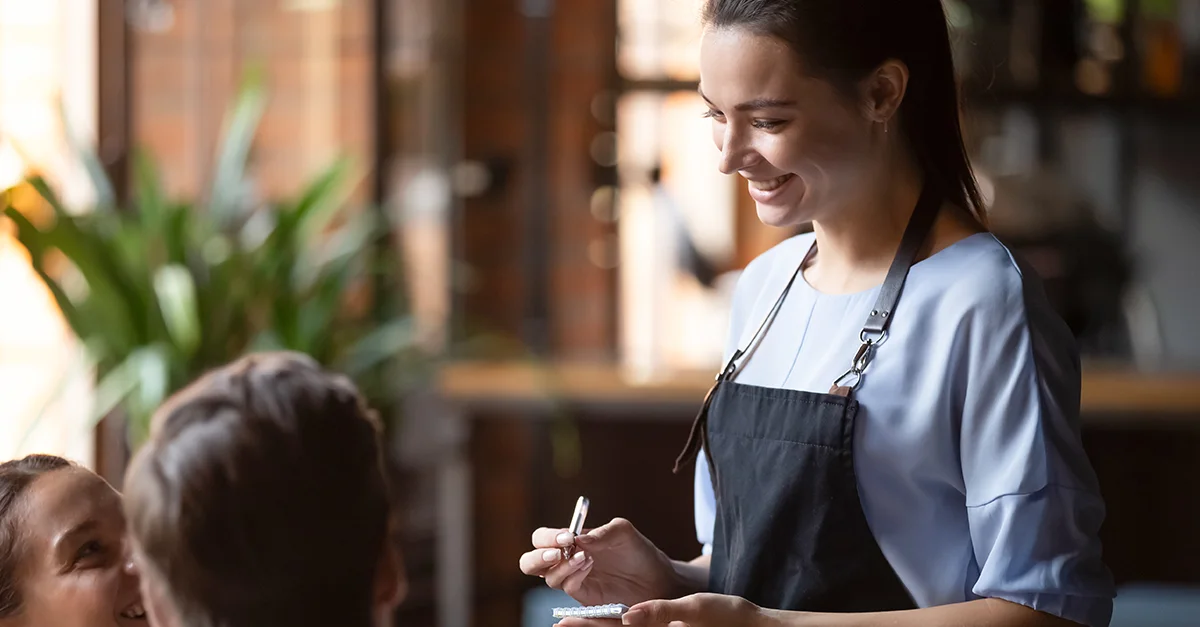 Café staff member wearing an apron, writing on a notepad while taking an order from a seated customer in a warmly lit coffee shop setting.