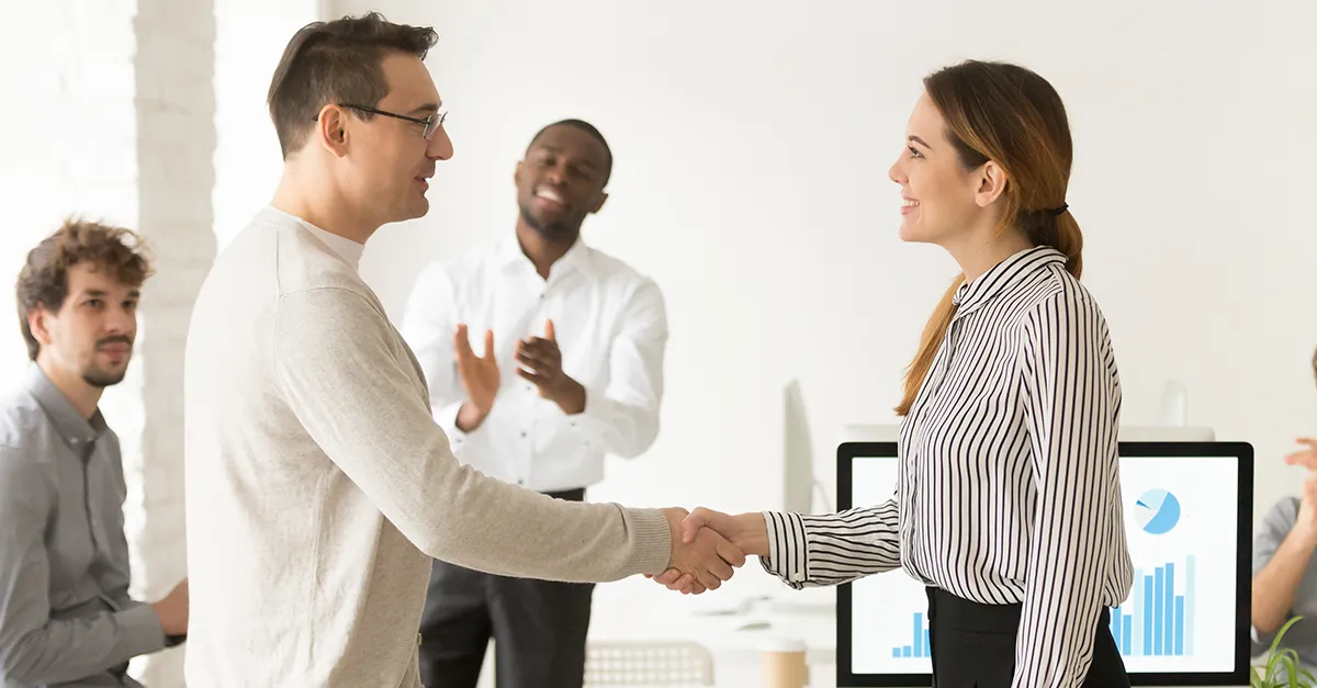 Two coworkers shaking hands in a bright office workspace after a meeting, with colleagues seated in the background and presentation screens visible.