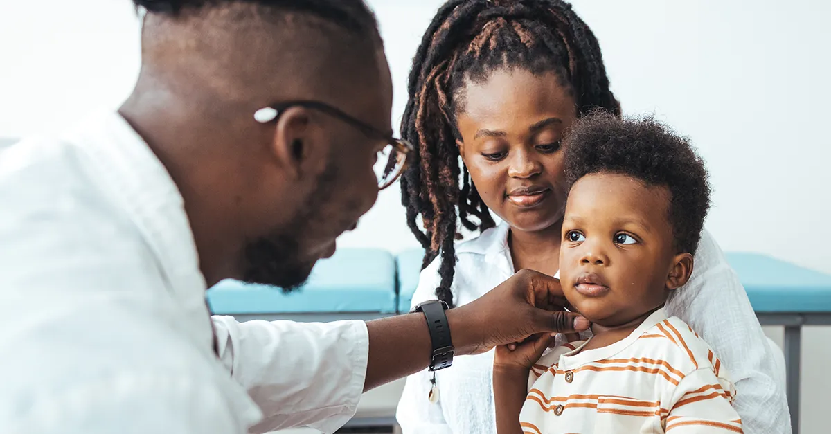 Healthcare professional using a medical instrument to examine a young child’s ear while an adult sits nearby, in a clinic or medical setting.