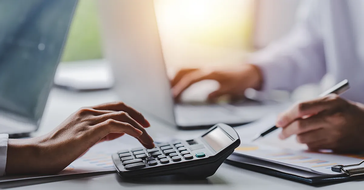 Close-up of two people at a desk, one using a calculator while another writes on a document, with a laptop and papers visible, representing financial analysis or budgeting tasks.