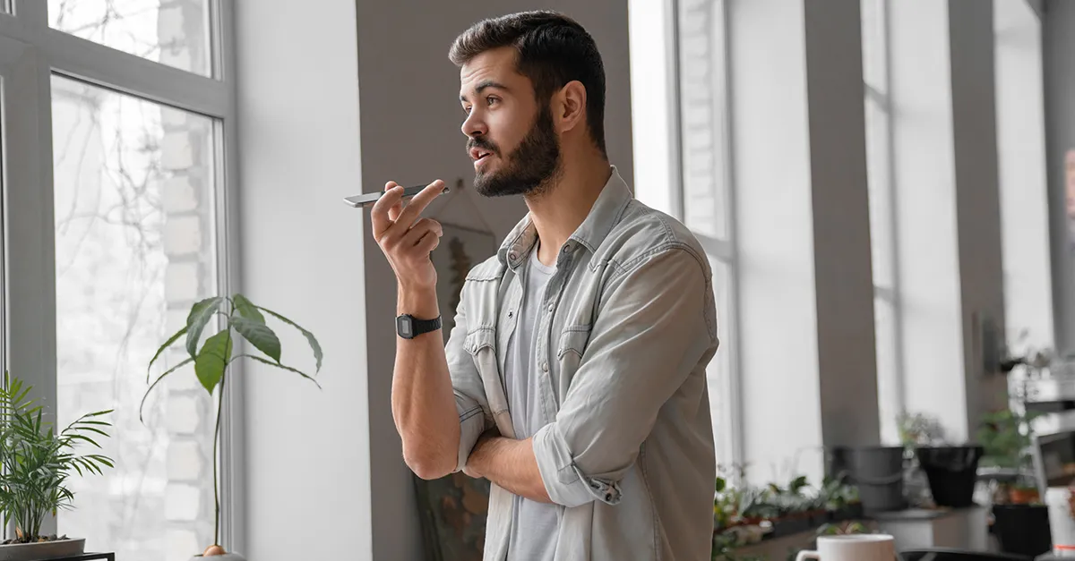 Person standing near a window indoors, holding a smartphone close to the mouth while speaking, with plants and natural light visible in a home setting.